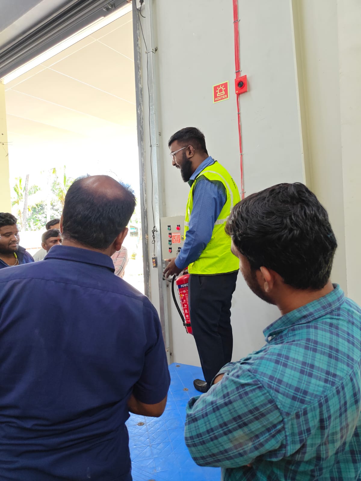 Site team inspecting a fire alarm hooter panel and extinguisher mounted on a shutter wall