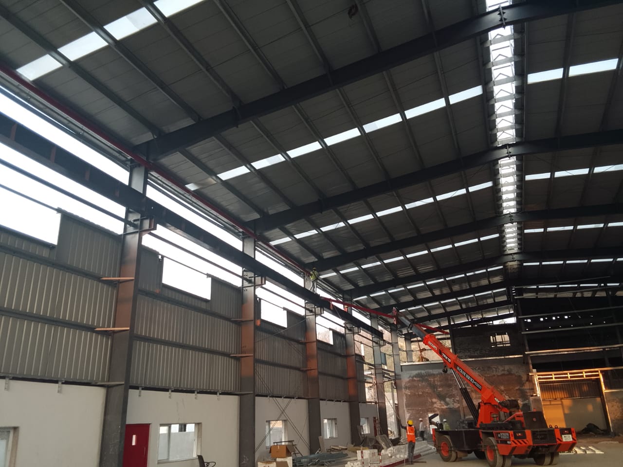 Interior of a steel-frame industrial shed under construction, workers routing fire piping along the roof trusses