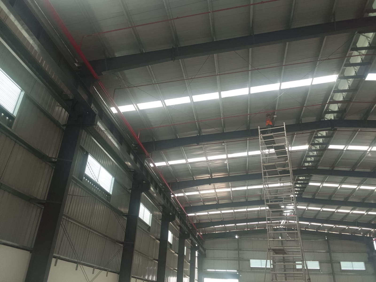 Industrial shed roof structure with a worker on a scissor lift installing fire piping near the trusses