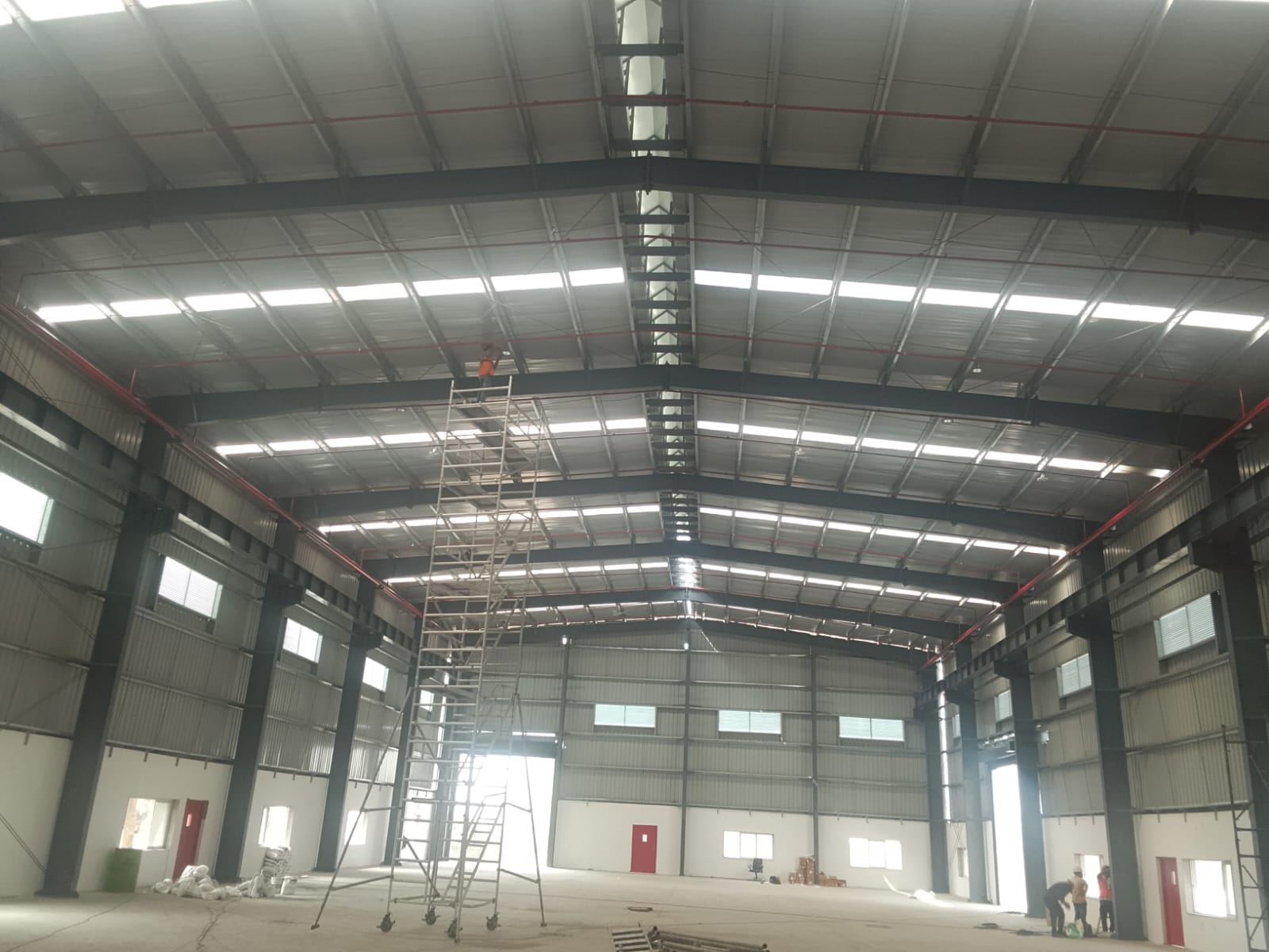 Wide interior view of an industrial shed under construction with a mobile scaffold tower and workers below